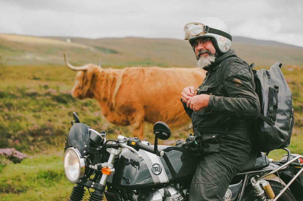 Scottish Motorcycling on Scottish West Coast. A motorcyclist riding a Royal Enfield GT Continental rides alongside a Highland Cow