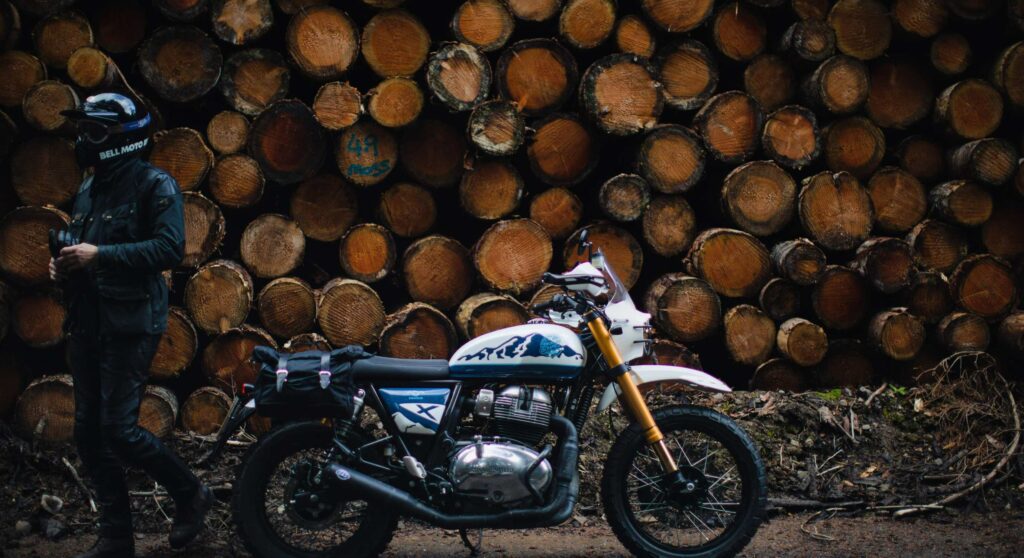 A Motorcyclist with custom Royal Enfield 650 stands next to wood stack in The Scottish Highlands
